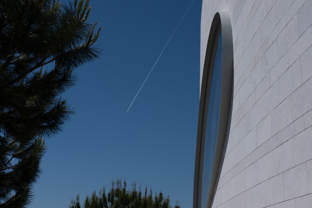 A detail shot of one oval window of the Champalimaud Foundation. On the right the window is shown at an acute angle, with deep blue sky behind and tree ferns encroaching. In the background a plane can be seen flying away with a white contrail visible against the deep blue sky.