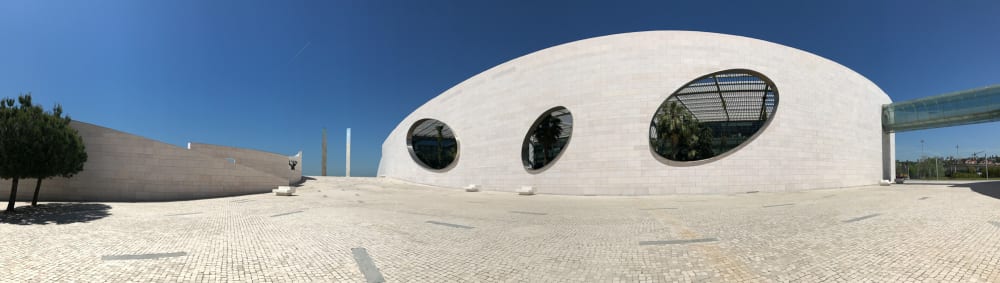 A panorama in bright sunlight of the Champalimaud Foundation courtyard. On the right the large main wall of the building is visible with three large oval openings cut away.