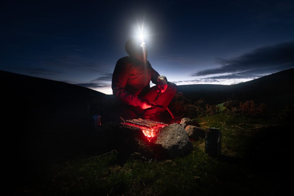 A figure is lit by glowing embers of a bbq at night. He’s wearing a head torch and looking to the sky. In the background, the last of the daylight is visible on the horizon.