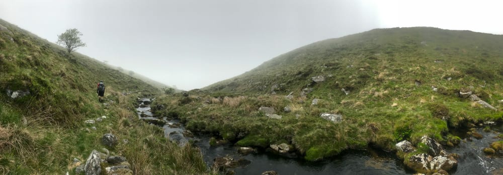 A panorama taking on a foggy day from the side of a small stream. A hiker is ahead on the stream walking downhill.