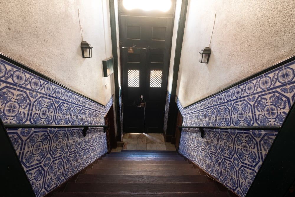 A wide angle photo looking down an interior staircase to the front door of a building. The staircase is steep, and on either side the walls are covered to shoulder height with blue and white patterned tiles.