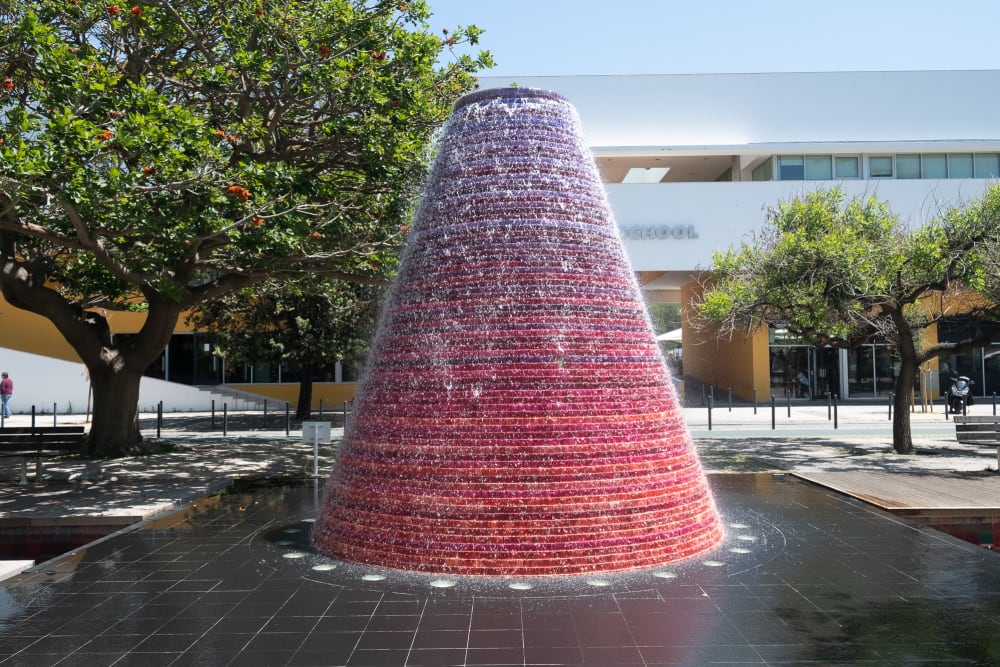 A large fountain in the centre of the frame on a sunny day. The fountain is made from a large circular cone shape, purple-red at the top, fading to red at the bottom.
