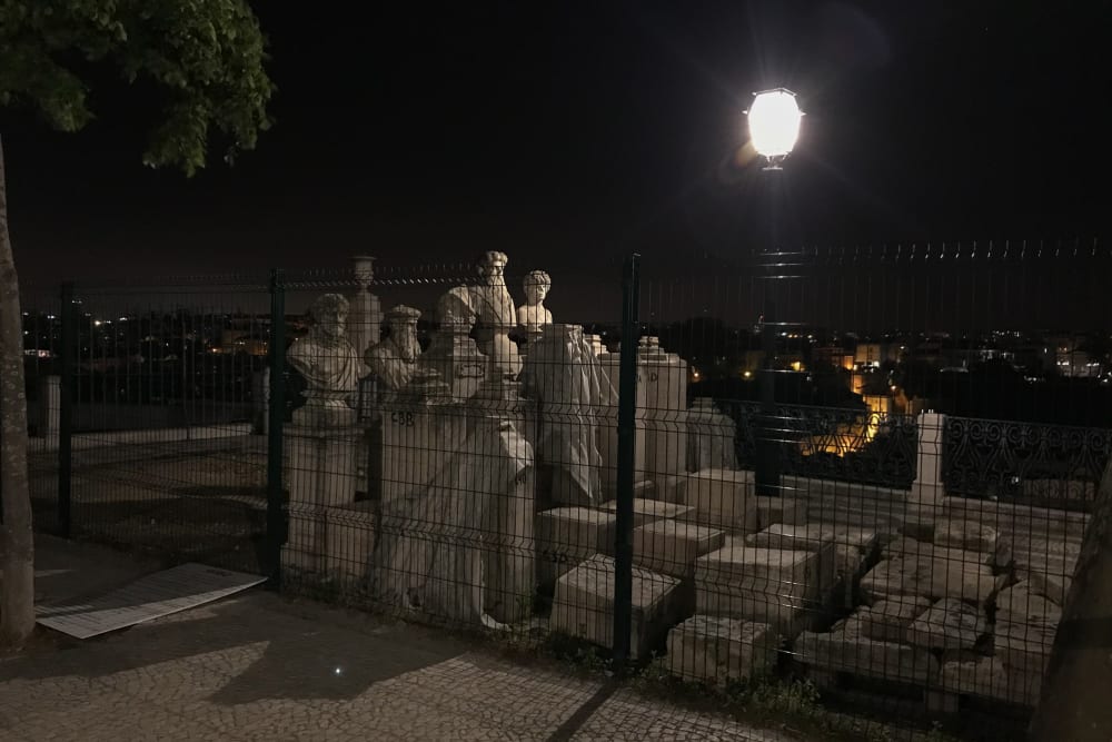 A group of tall marble statues are arranged in a tight grid and surrounded by tall fencing. They’re lit from one side by a streetlamp.