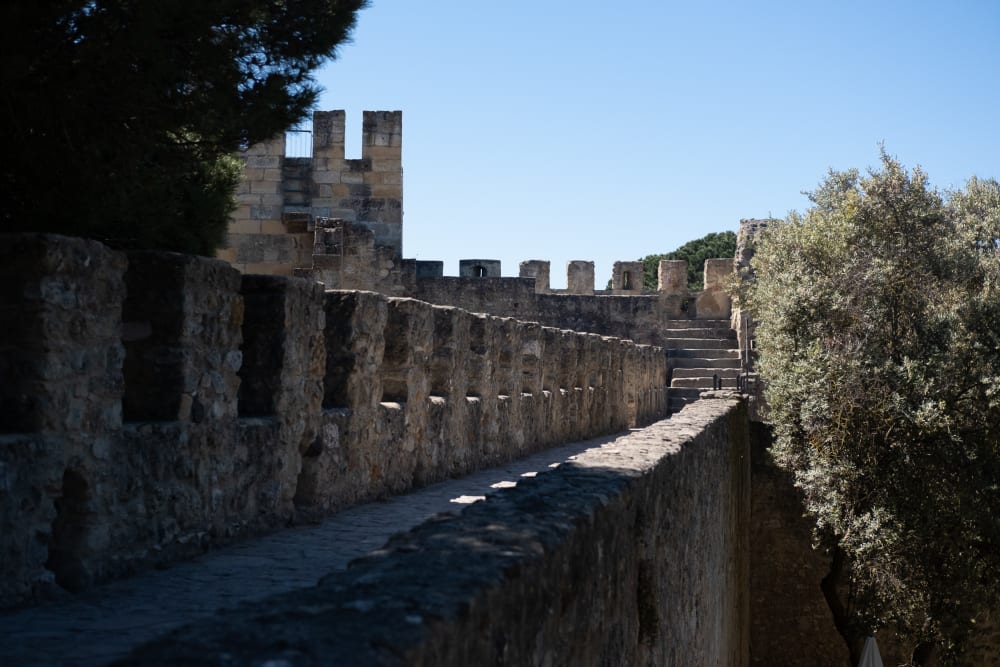 Looking down a series of regular castle battlements. A large olive tree as tall as the battlements is visible on the right.