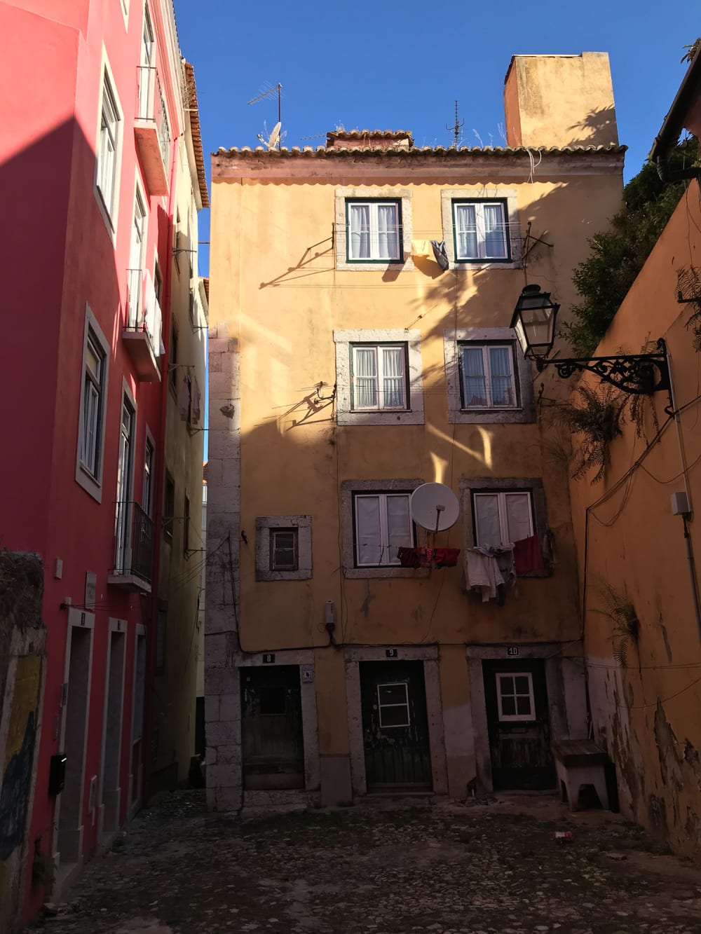 A portrait photo of the entrance to a 4 storey building at the end of a wide alleyway. The building is painted in pastel-yellow, and has a pastel pink building to the left.