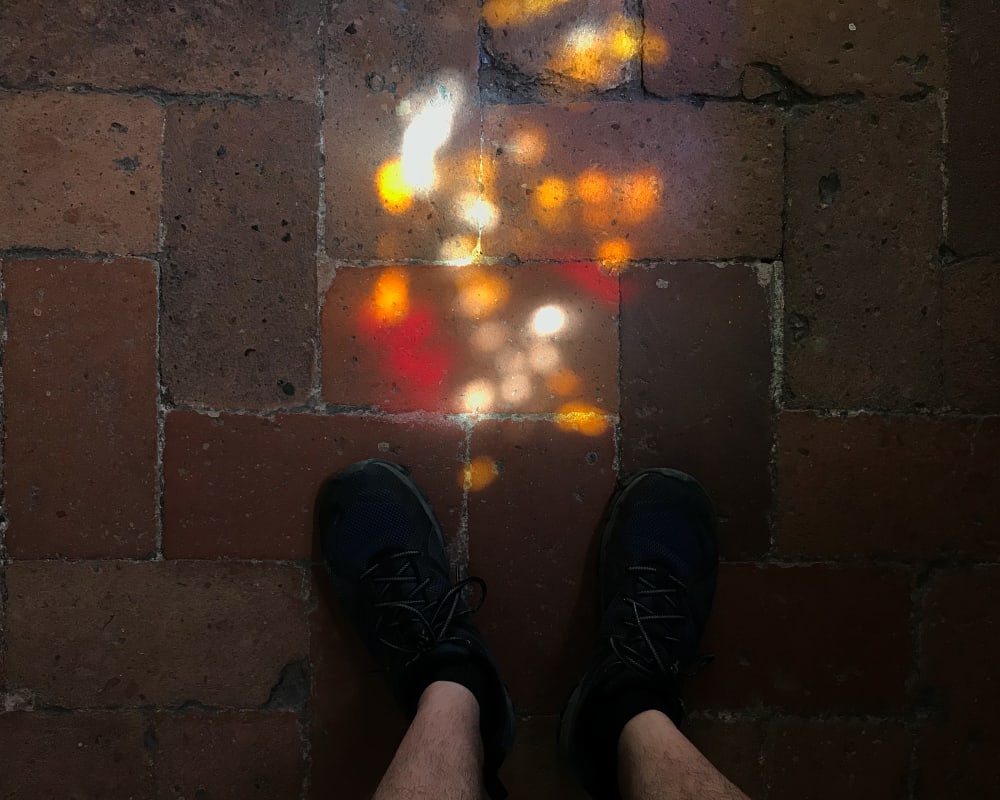 Looking down at a parque brick floor. At the base of the image are the photographer’s feet. Above them is speckled bright dots of light.