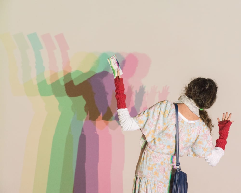 A woman stands in front of a white wall. A light shines on her from behind, casting a multi-coloured shadow on the wall.