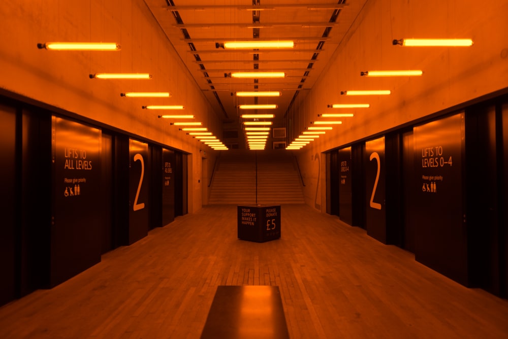 Looking straight on to the elevator lobby in Tate Modern. The room is filled with orange light from a series of regularly spaced fluorescent lamps.