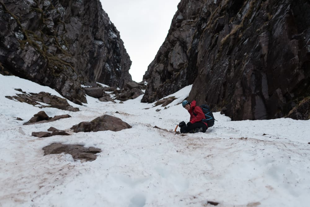 Looking up a snowy gully. Chris is mid way up the frame, crouched in the snow, looking at the camera.