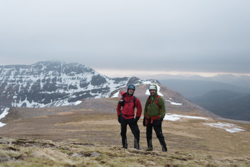 Chris and Ed pose for a photo on top of Beinn Eighe. They both have heavy jackets and helmets on.