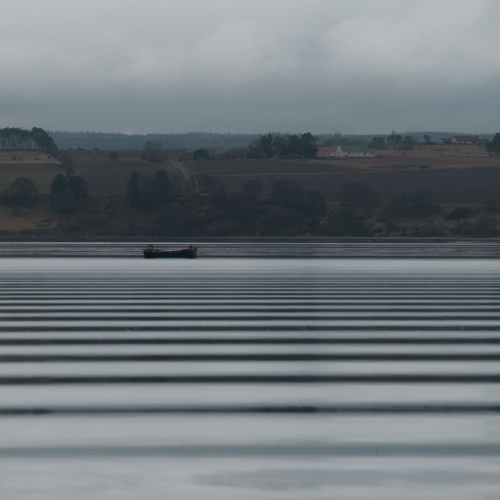 Looking across a wide lake on a grey day. In the distance is a small industrial boat. The water has regular horizontal dark bands - like a barcode - caused by ripples from a passing boat.