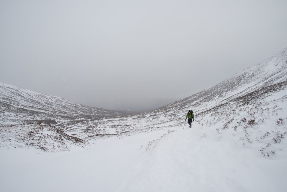 looking down a snowy hill with low grey clouds. Ed is off to one side walking up towards the camera.