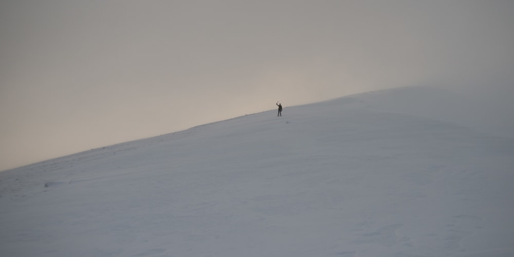 A wide shot at dusk looking up a snowy hill. A figure (Ed) in the distance can be seen waving an ice axe.