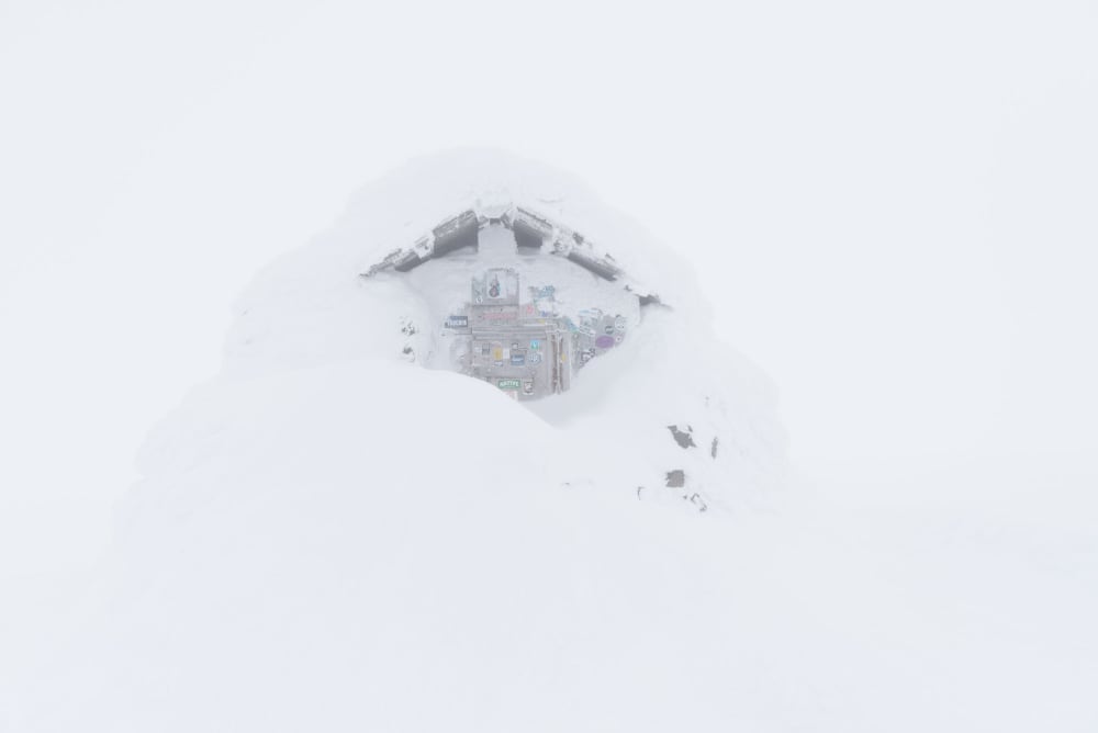 The emergency hut at the top of Ben Nevis. It’s covered in lots of snow, so only a patch of the top of the doorway is visible. There’s low visibility so it looks somewhat ghostly.