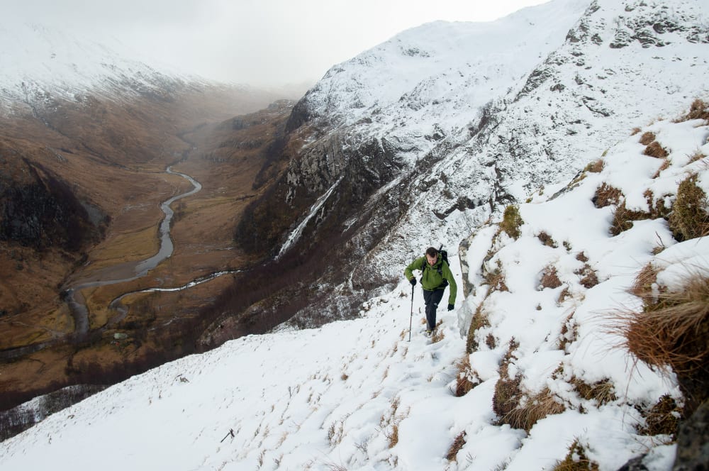 Ed climbing a snowy and grassy mountainside. In the far distance the brown base of the valley can be seen with a river running through the middle.