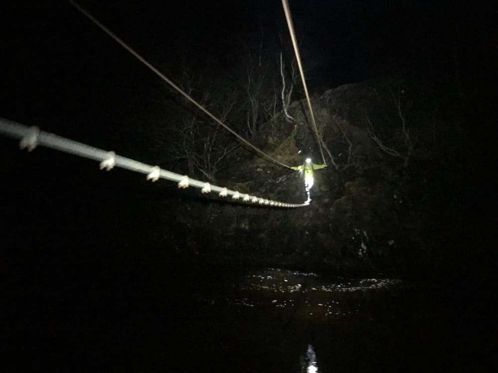 Chris crossing a rope bridge over a river, coming towards the camera. It’s pitch black, and he’s got a headtorch to lead the way.