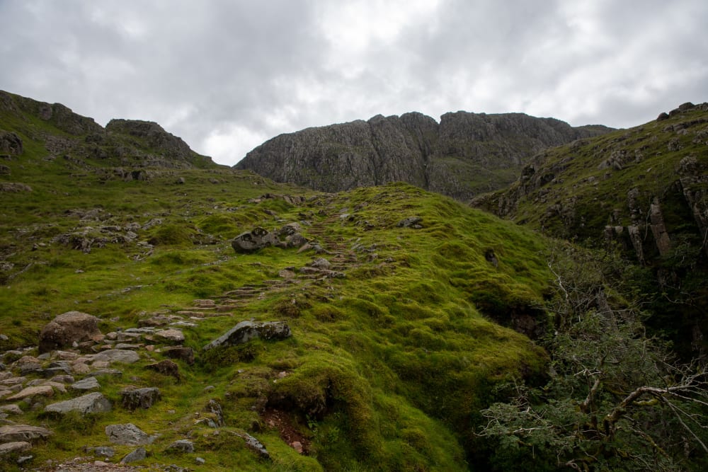 A dirt path with some steps winds up a dark green moss and grass covered hill, leading to a large rock formation in the distance.