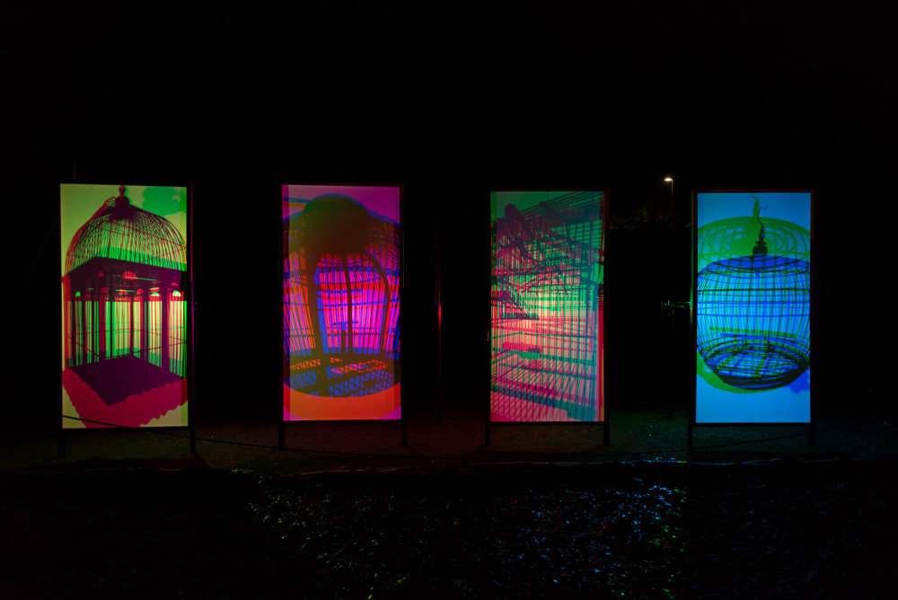 Four vertical panels are side by side in the dark. They’re light from behind with bright neon colours, projected with shadows of birdcages.