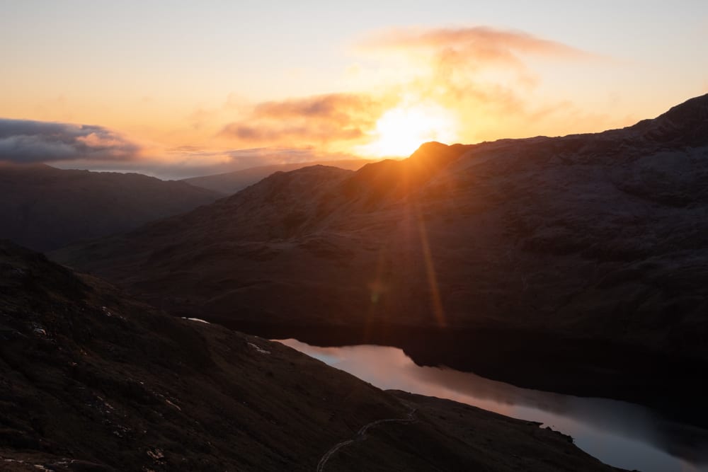 A sunrise photo taken from midway up Snowdon, facing the sun. There’s a still lake on the bottom, reflecting some of the sunrise.