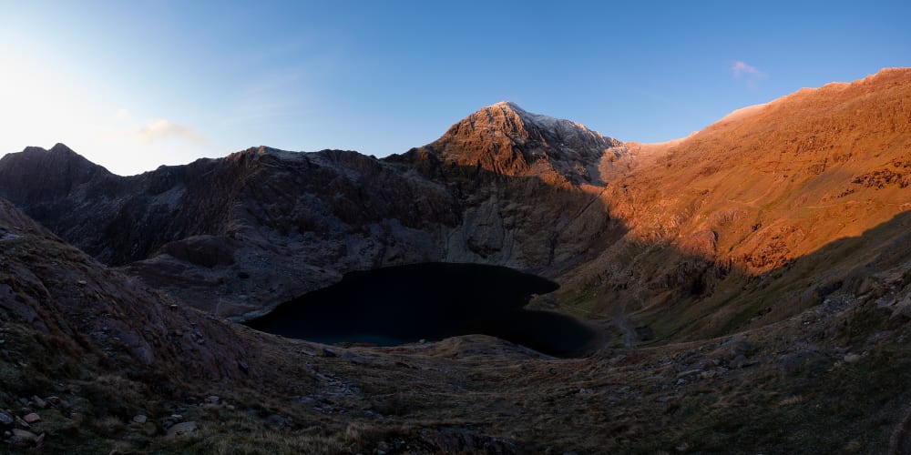 A panorama of the bowl of Snowdon taken just after sunrise. The right side of the mountain is bathed in orange sunlight, whilst the bowl and left are still in shadow.