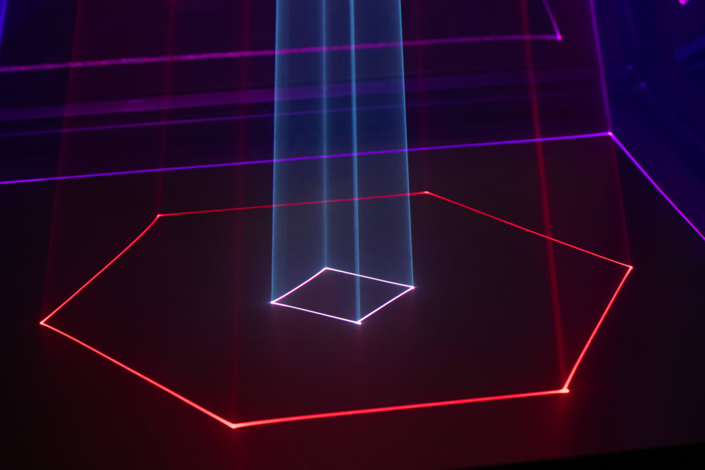 A laser beam projects shapes from above on to the ground. It has traced a large red hexagon, with a small pink square inside.