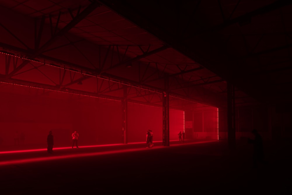 A photo in a large dark warehouse. The only light is deep red in one section of the warehouse. Several still figures can be seen around.