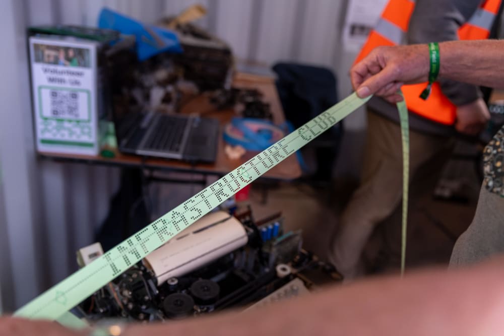 A long strip of paper is held up between two hands, with holes punched along the length spelling the words ‘infrastructure club’.