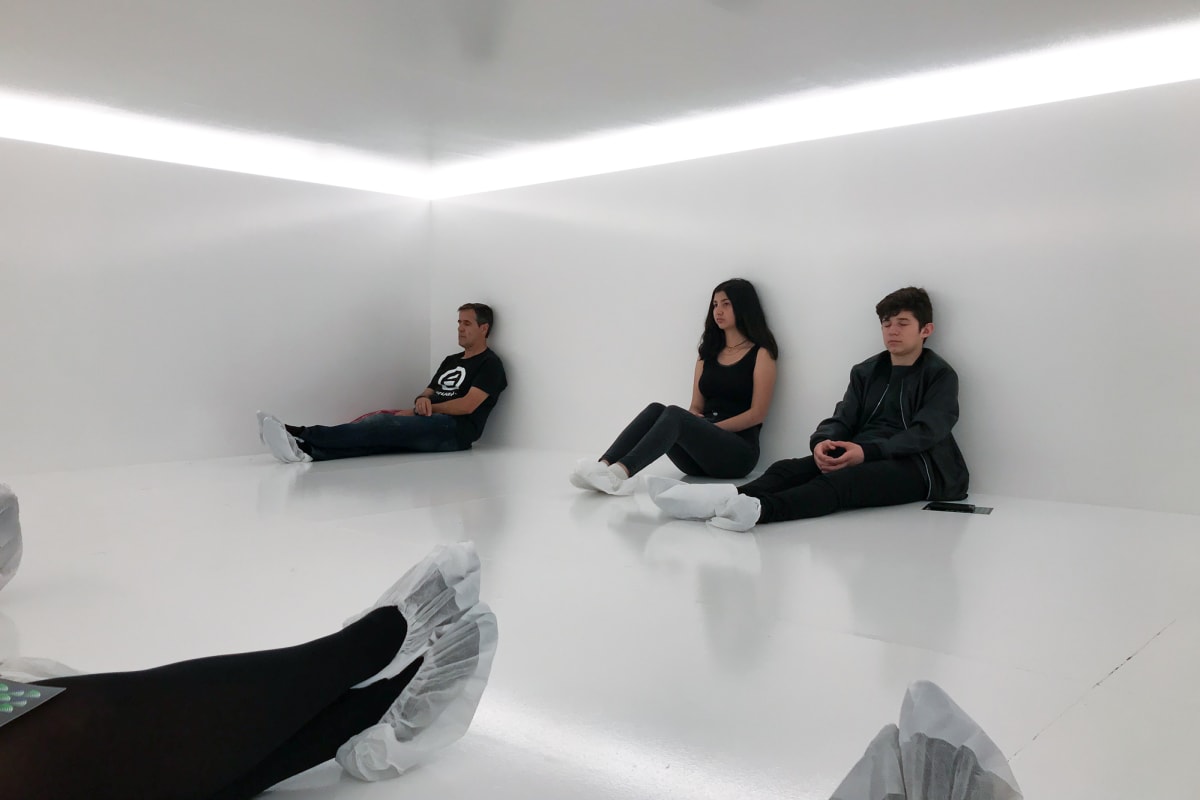 Several people with paper socks are sat in a gloss white room with white light overhead.