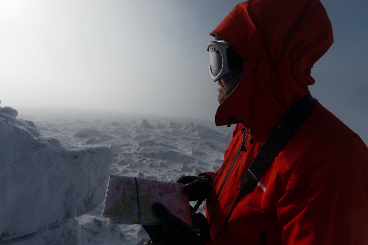 Ed looks out in to the distance, which is misty with low visibility. He’s holding a map and compass, and wearing a bright orange jacket and ski goggles.