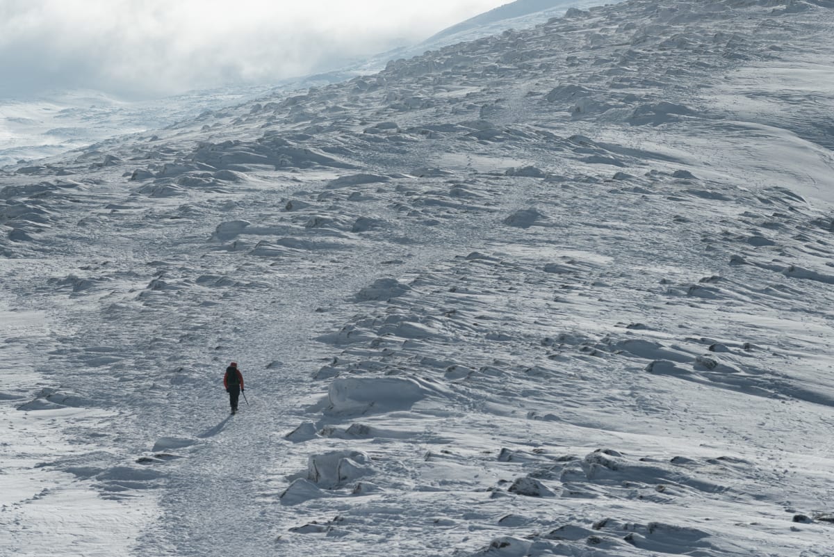 A telephoto photo of Ed walking up a wide snowy hill. He’s a small dark figure in an otherwise very bleak landscape. There’s an ice axe in his right hand.