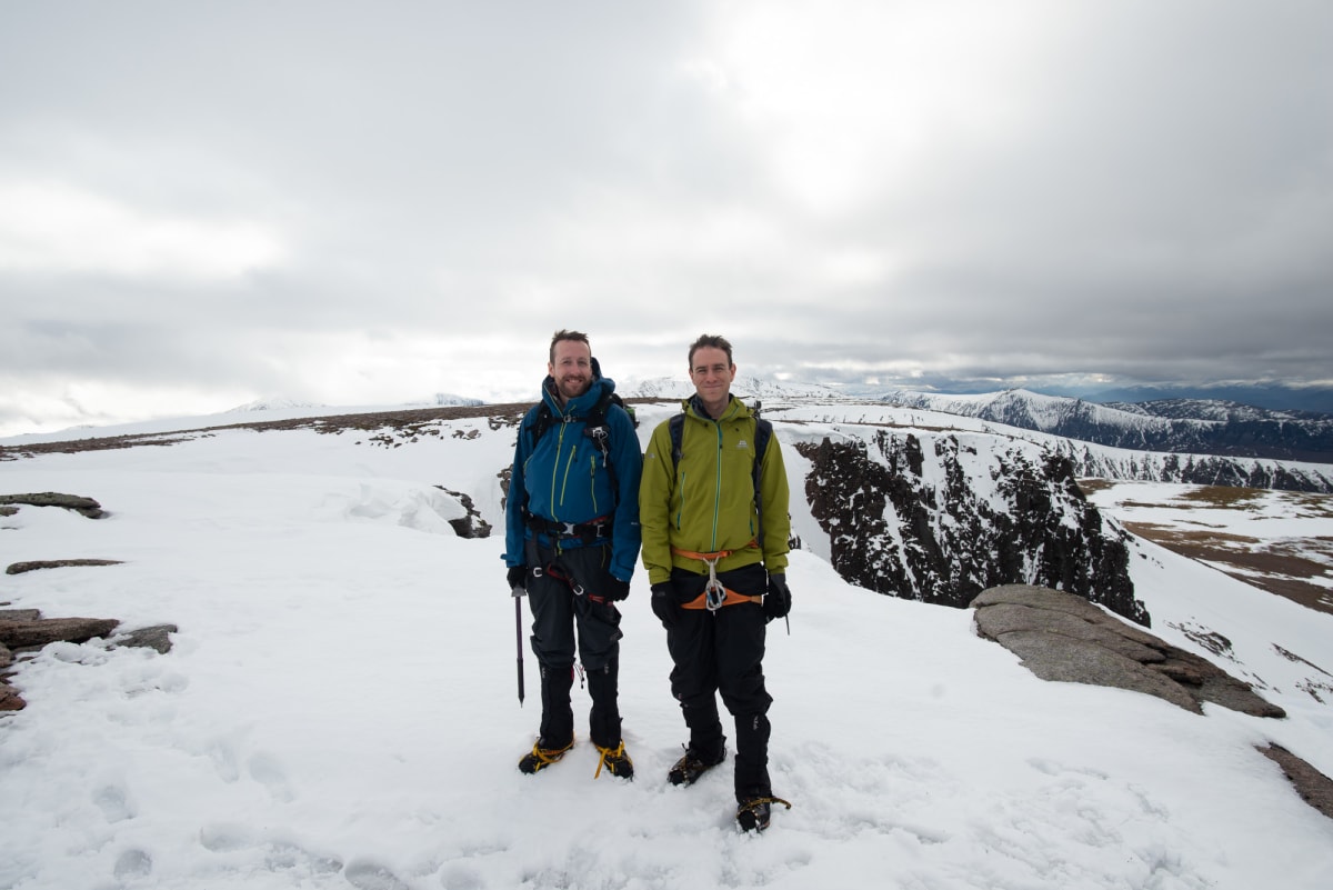 Chris and Ed stand at the top of Fiacaill ridge. The ground is mostly snowy with patches of brown.