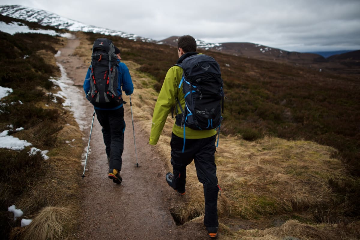 Chris and our instructor walk along a path uphill. There’s small patches of snow in the distance, but the ground is otherwise brown and windswept.