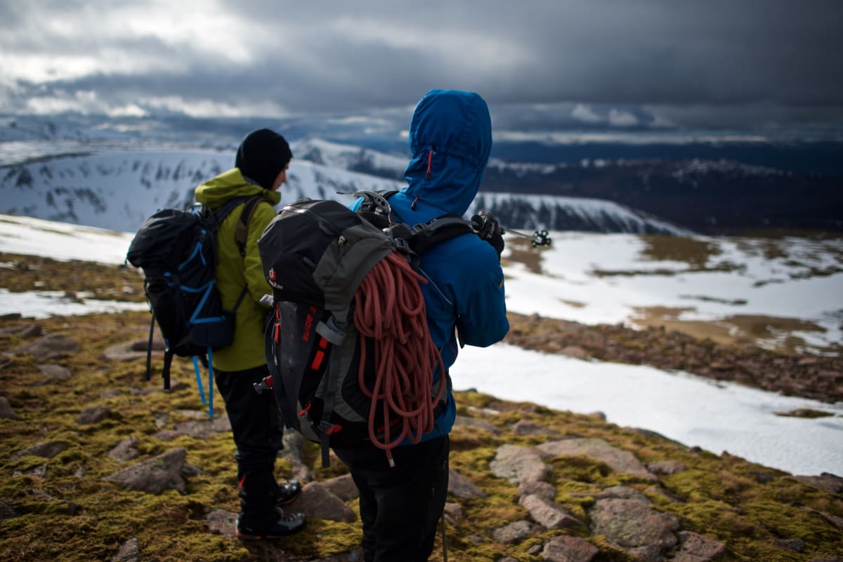 Photo of the backs of Chris and our instructor. Our instructor is pointing downhill with a ski pole. There’s dramatic lighting in the sky - sunlight in the foreground and ominous dark clouds in the background. There are patches of snow on the ground.