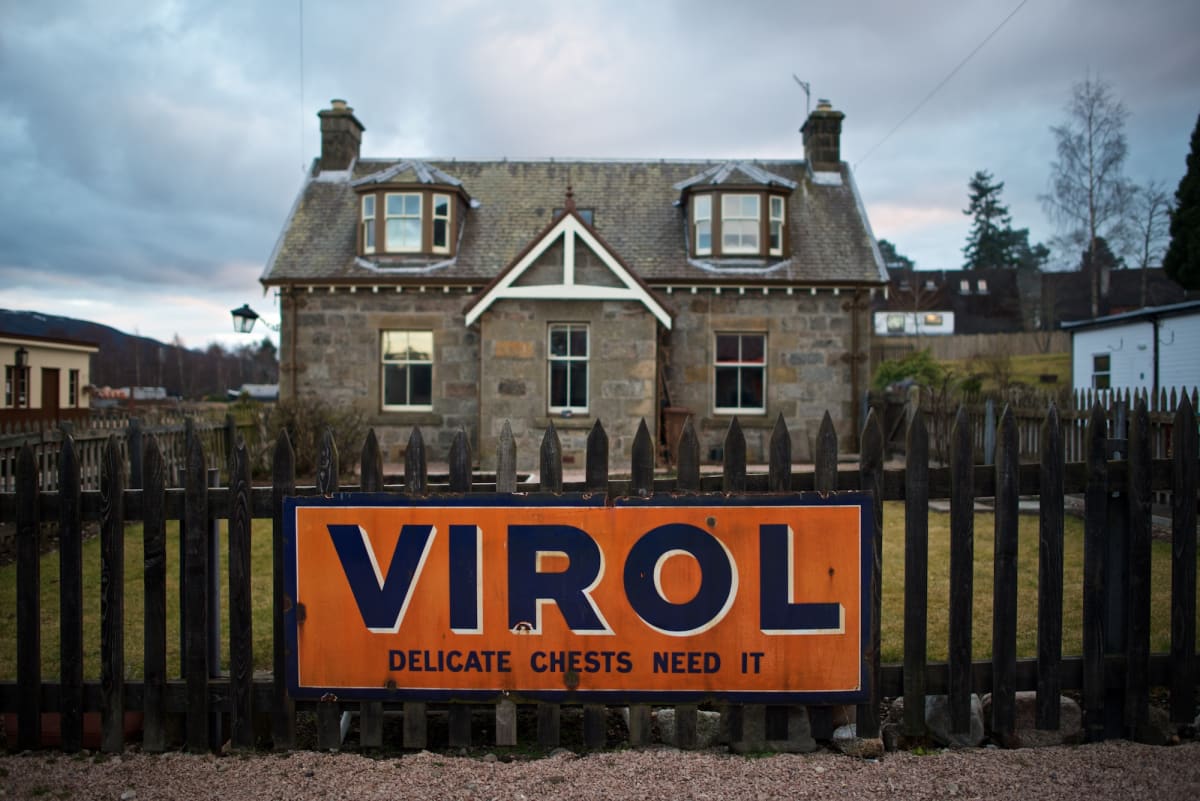 Facing straight on to a large orange sign in front of a cottage. The sign is written in large uppercase blue letters and reads ‘VIROL’.
