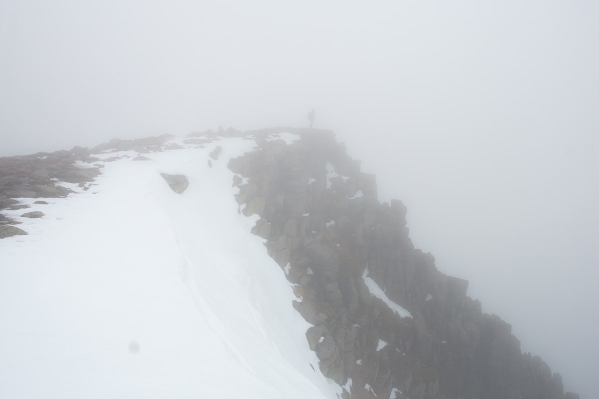 A photo of a rocky cliff partially covered in snow. There’s strong fog everywhere so the background and bottom of the cliff can’t be seen. There’s a shadow of a person at the tip of the cliff.