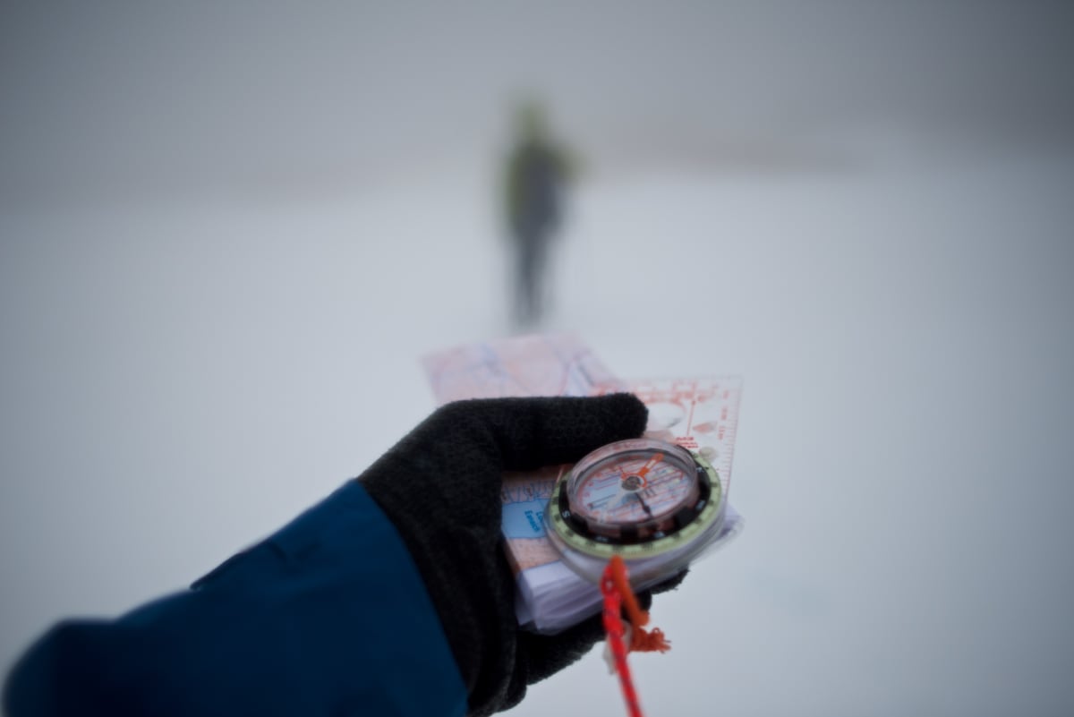 A gloved hand holds a compass and map in front of the camera as a blurred figure walks ahead in the distance surrounded by snow and fog.