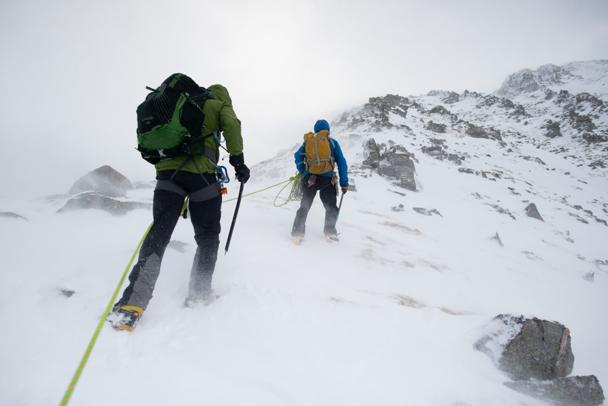 Ed and a guide walk up a snowy mountain on an overcast day. The camera angle is near to the ground looking up at their backs. They’re both connected by a climbing rope.