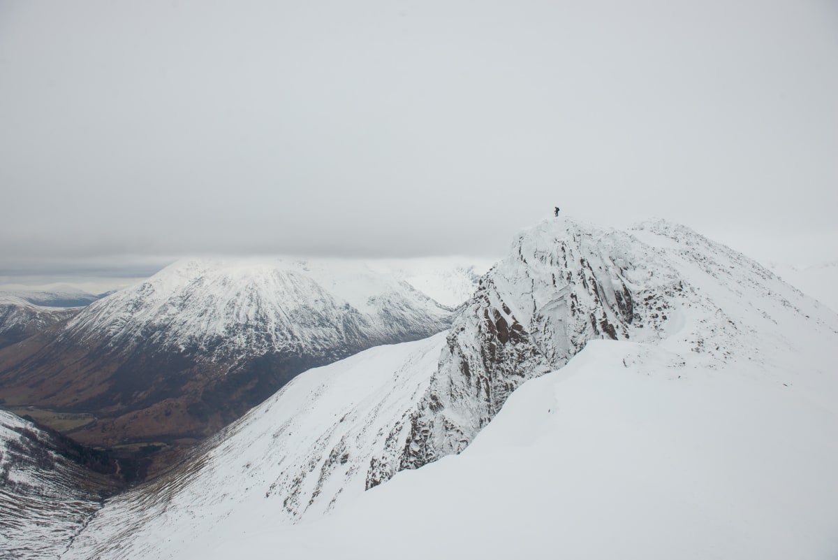 A wide photo of a snow covered mountain peak with other mountains in the far distance beyond a valley. At the top of the peak is a small figure.