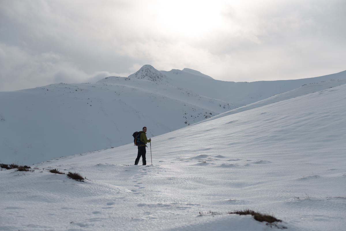 Chris stands at the foot of a snowy coire with early morning sun ahead of him.