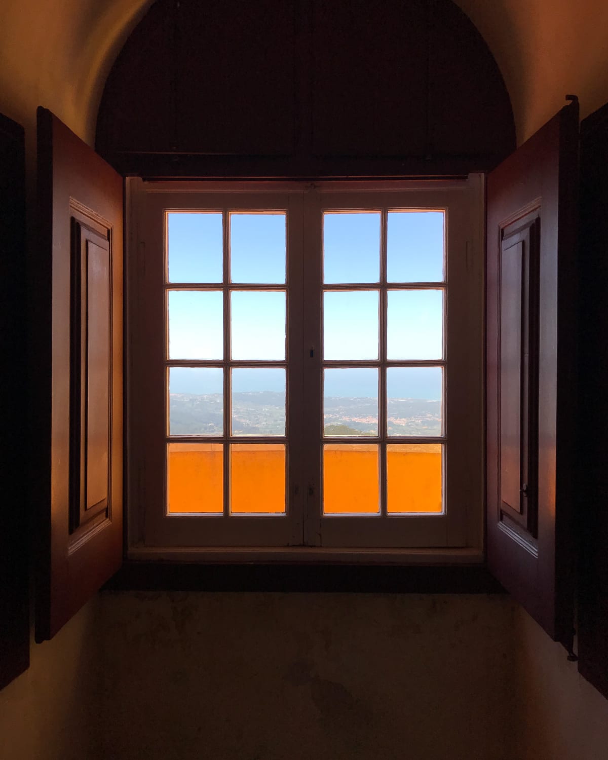 Looking from inside to a window looking out on a high vista. The interior is dark with dark wood window frame. Through the window the sky outside is bright. The window is made from two sets of 2x4 small glass frames. The bottom row is bright orange from an balcony outside.