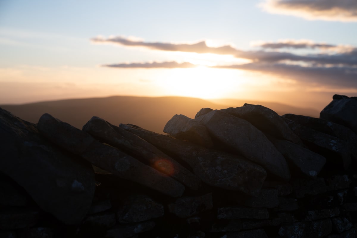 A close photo of the top row of stones of a stone all. The stones all lean to one side. In the background the sun is just setting.