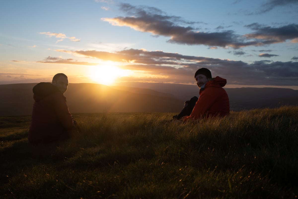 Ed and Chris sit on long grass in late golden sunset.