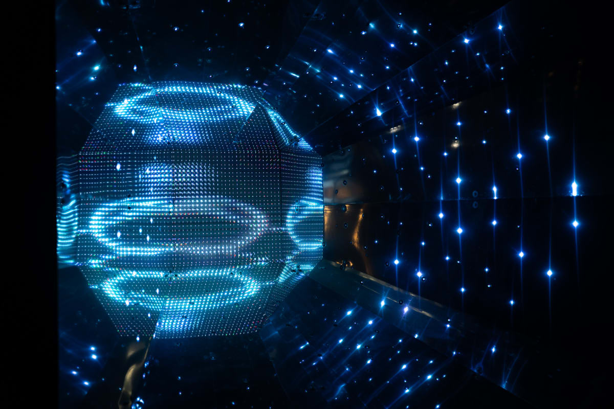 A dark photo with ball shaped object on the left and surrounded by dots of light like stars. The object is similar to a mirror ball but with flat faces and lit up like an old CRT TV.