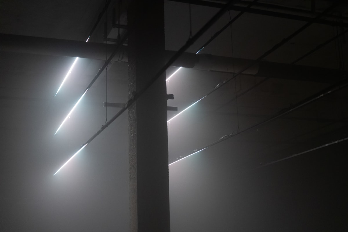 A dark scene with a single concrete pillar in the middle left. Six lines of light are evenly spaced vertically either side of the pillar. There’s fog in the room. Cables extend to and from the lines of light - reminiscent of telegraph poles.