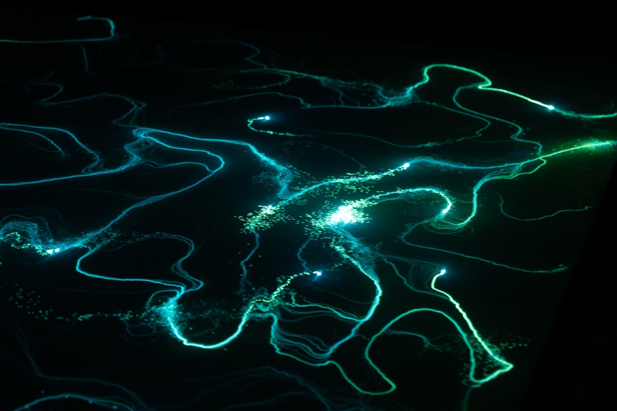 A photo from a moving light installation. A bright point light is projected on the floor, and wanders randomly around. The floor is covered in a material that glows for a period when exposed to light. The end effect is of random lines of light green and blue drawn all over the ground.