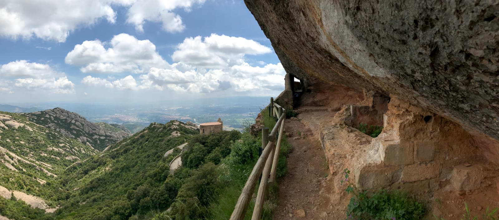 A panorama half looking over the Montserrat countryside and half looking at a path along the side of a mountain that is covered by steep rock.