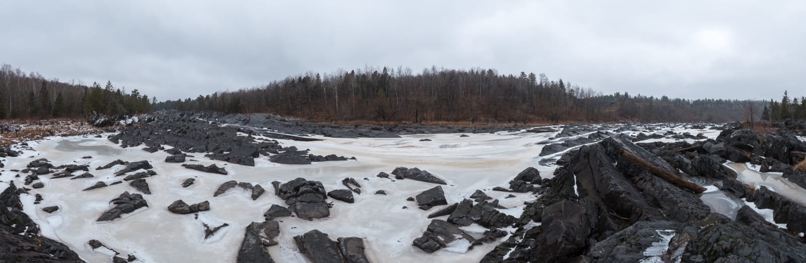 A panorama from the banks of an icy river. There’s dark stones littering the riverbed and creamy ice in between.