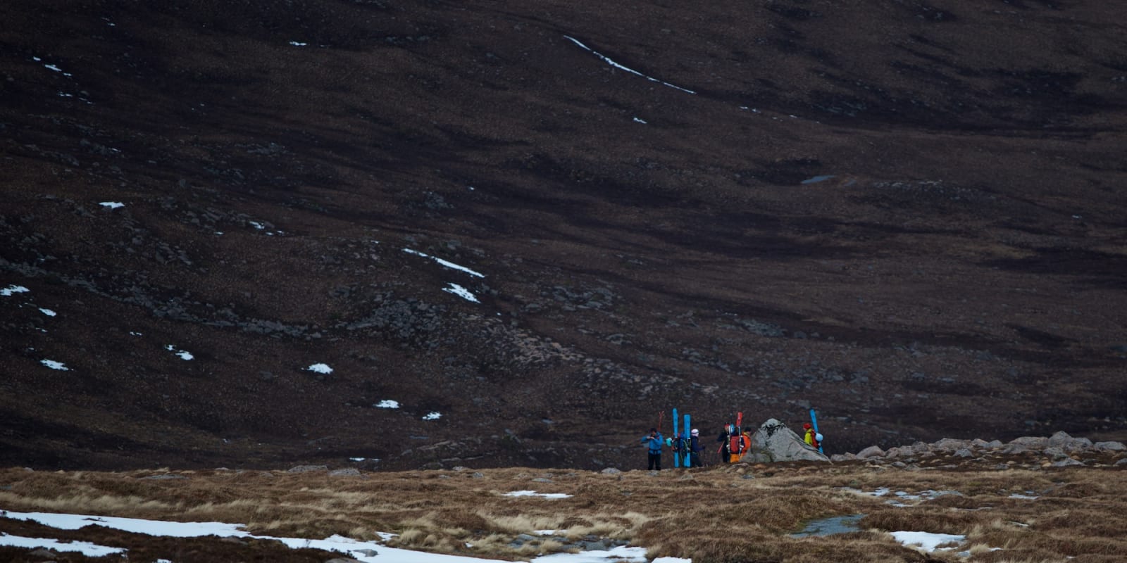 Long shot of several ski mountaineers in a valley. They have skied to the end of the snow, and are now packing up their gear.
