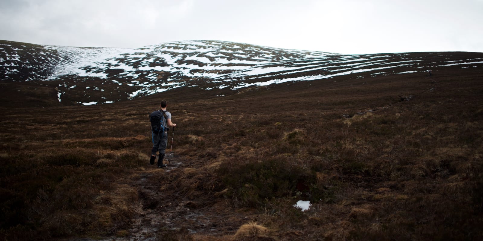 Chris hiking in a t-shirt on a brown grassy path. There’s partially snow covered hills in the distance.