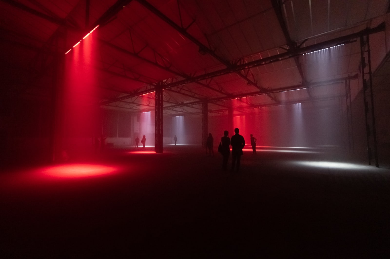 A cavernous dark warehouse. Several lights can be seen pointed at the floor like spotlights - some red and some white. Several figures can be seen silhouetted in the dark.